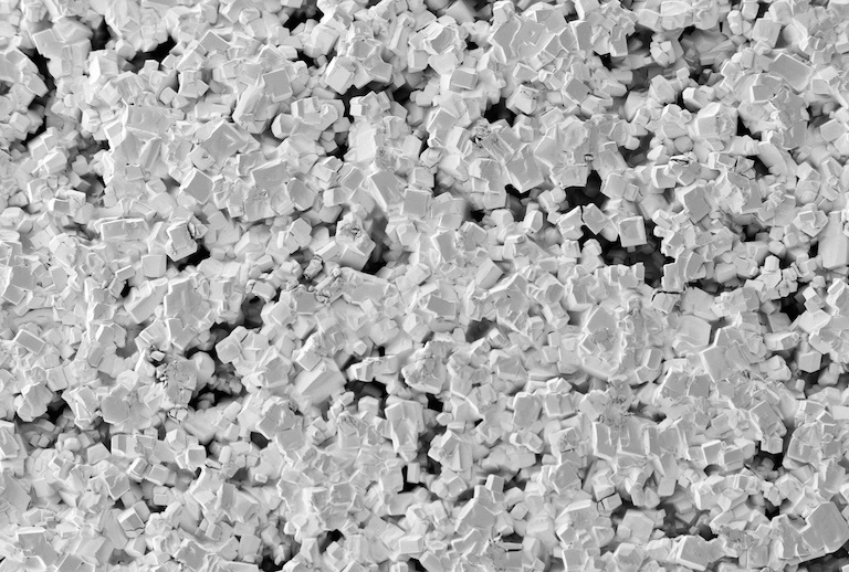 Black and white image of a pile of small square crystals. Appearing like square sponges pressed against glass. 